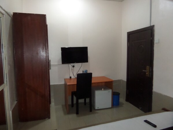 A small room with a wooden desk, a chair, a television mounted on the wall, a small cabinet, and a door.