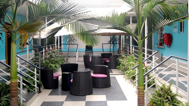 A spacious indoor area with a checkered floor, black wicker furniture with pink cushions, and tall palm trees. - Primal Hotel Ikeja GRA (Lagos)