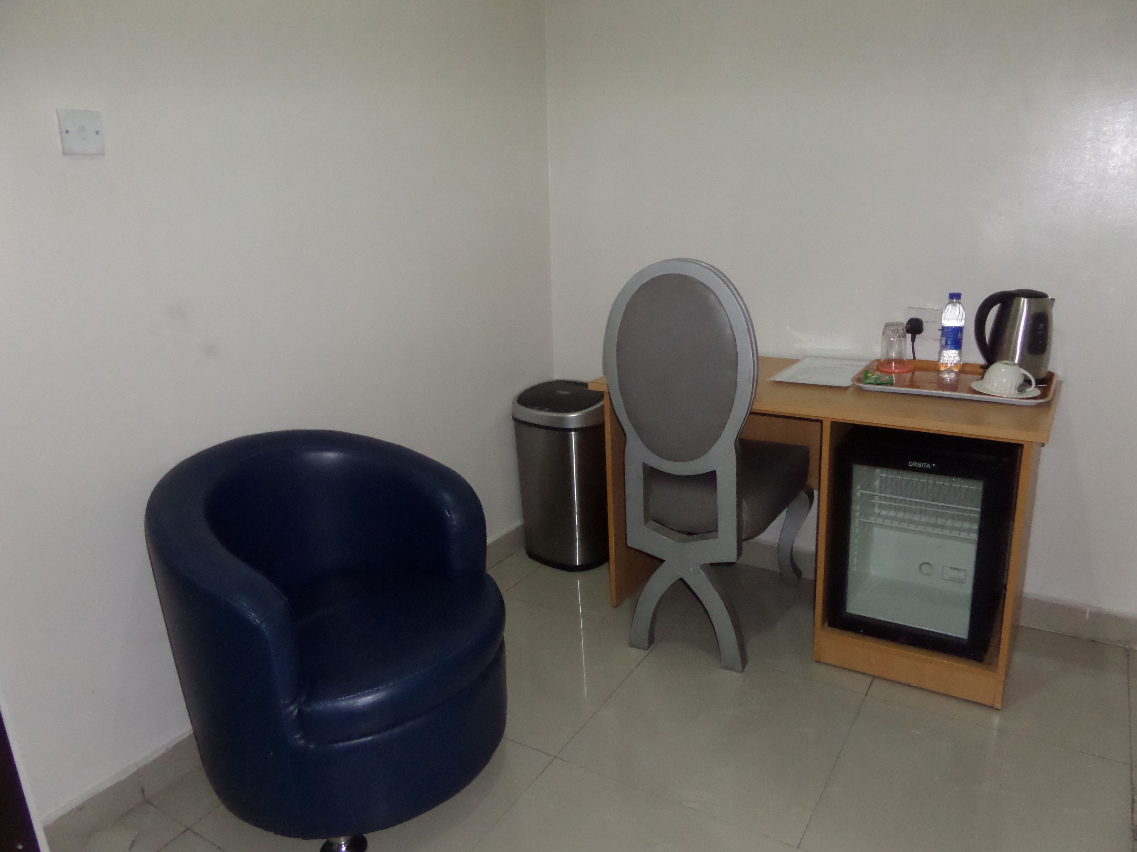 A small room with a blue chair, a desk with a computer, a trash bin, and a refrigerator.