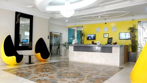 A reception area with a yellow wall, a reception desk, two black and yellow chairs, a large mirror, and a patterned floor. - Primal Hotel Ikeja GRA (Lagos)