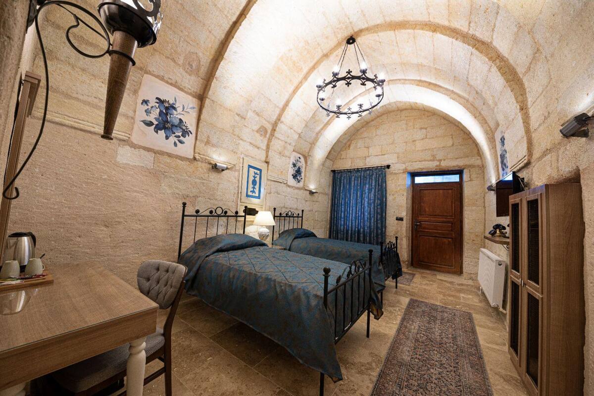 A room with two beds, a table with a kettle and cups, a chair, a lamp, and a wooden door.