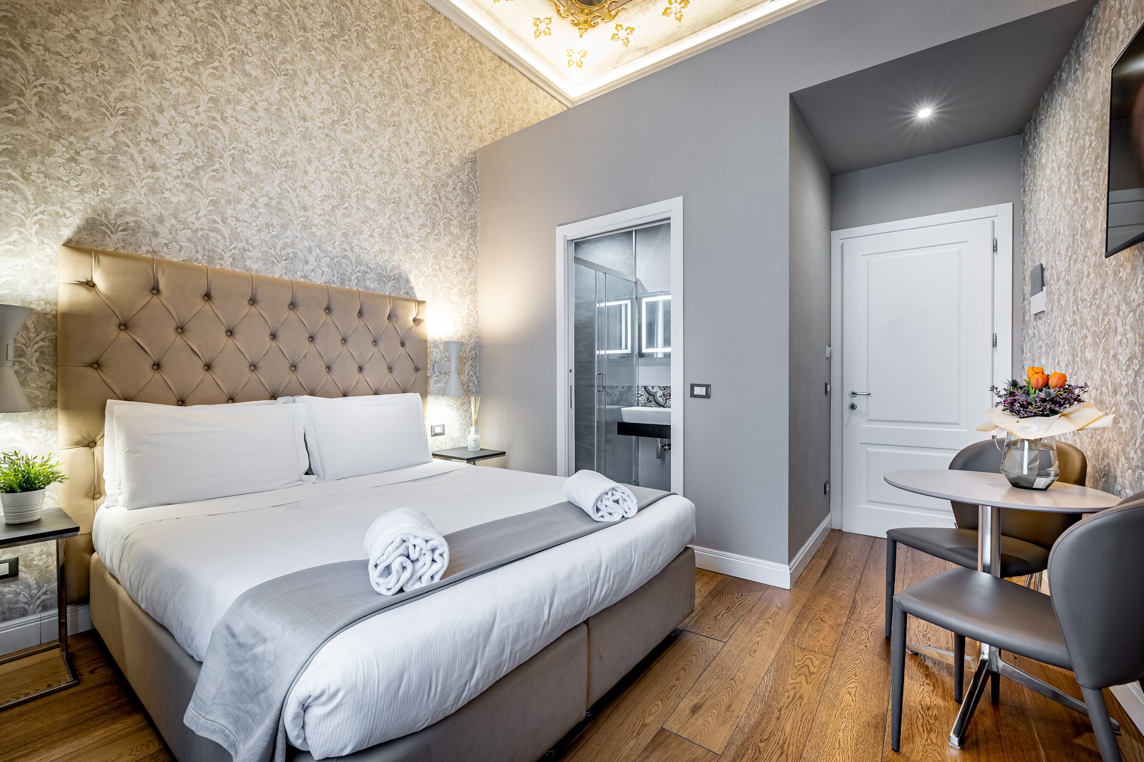 Quarto moderno com cabeceira estofada, cama grande, mesa de cabeceira com abajur e uma mesinha com um vaso de flores.