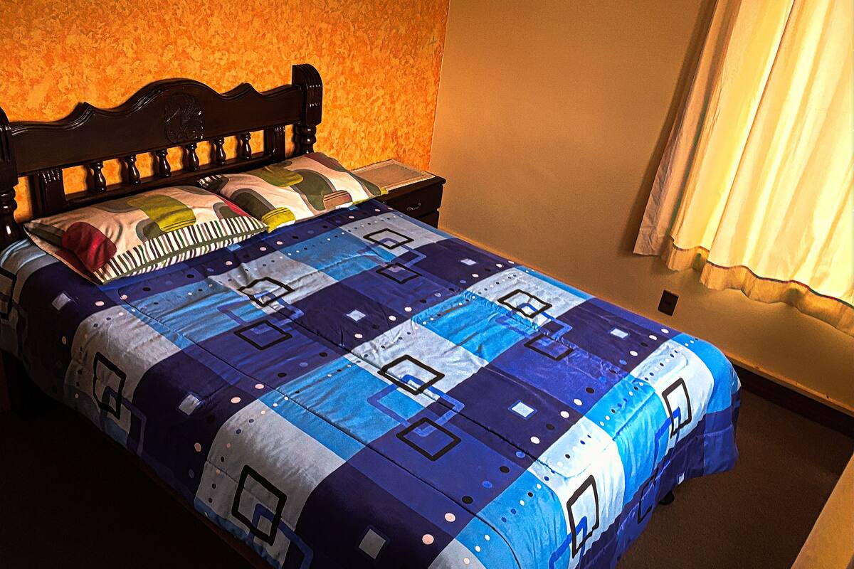 Ein Bett mit einer blau-weißen Steude, einem Holz-Kopfteil und einer schlichten Wand mit einem Vorhang.