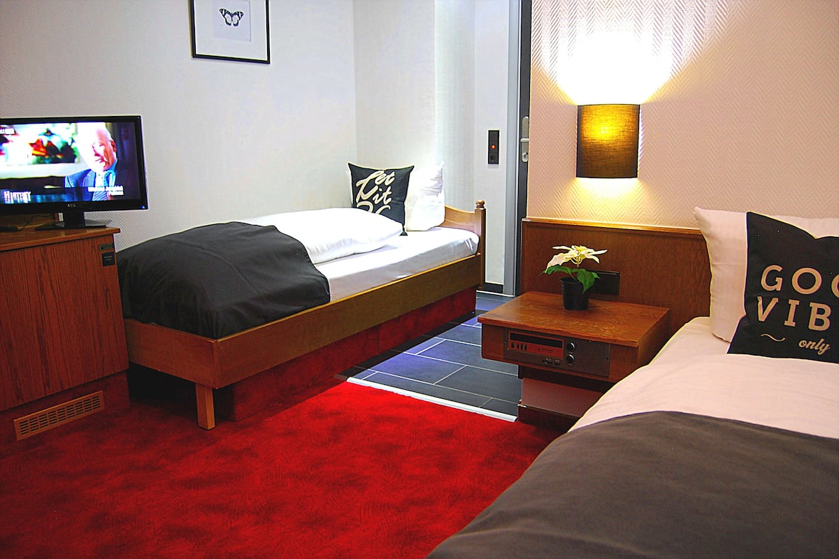 A hotel room with a bed, a bedside table with a lamp, a TV on a stand, and a red carpet.