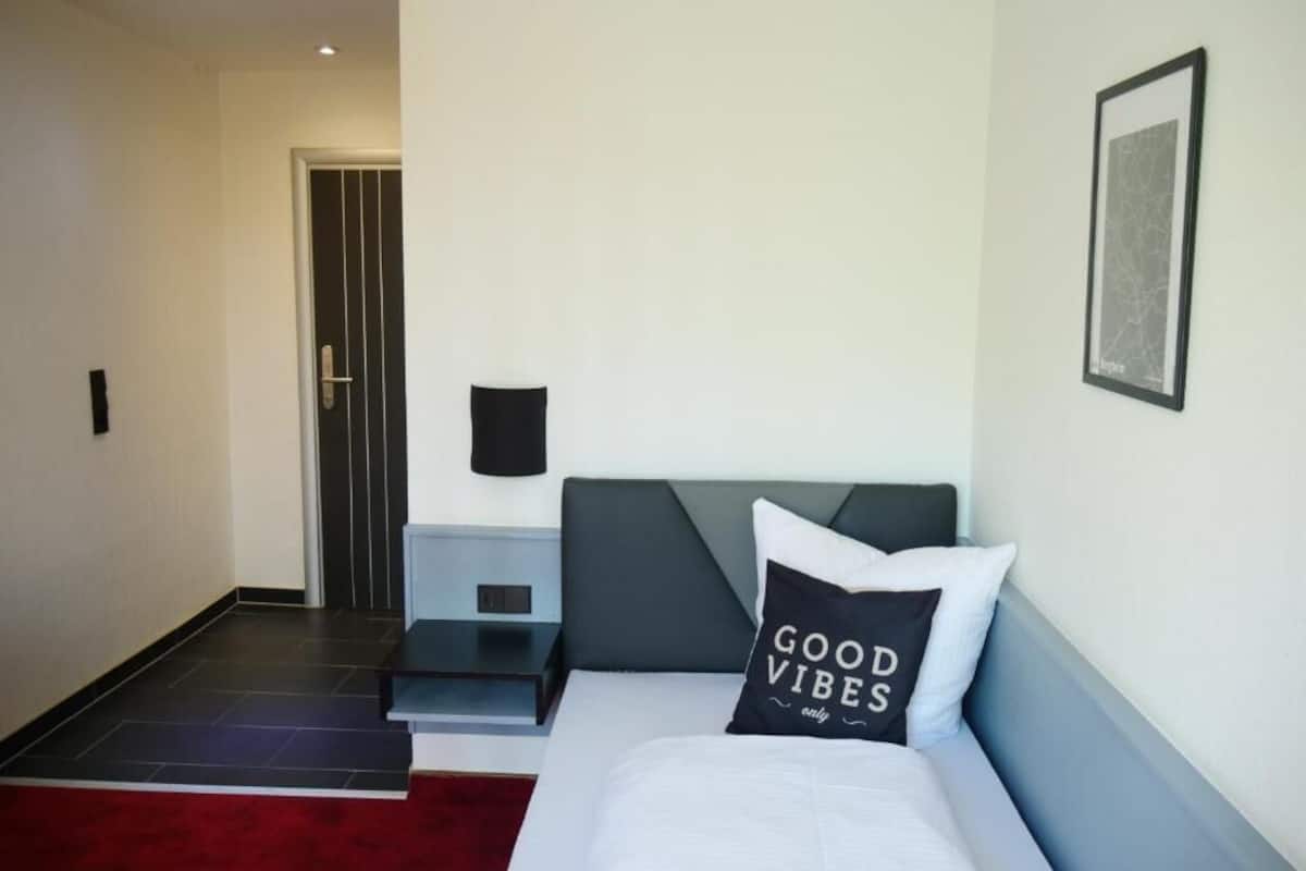 A hotel room with a bed, a nightstand, a wall-mounted lamp, and a door in the background.