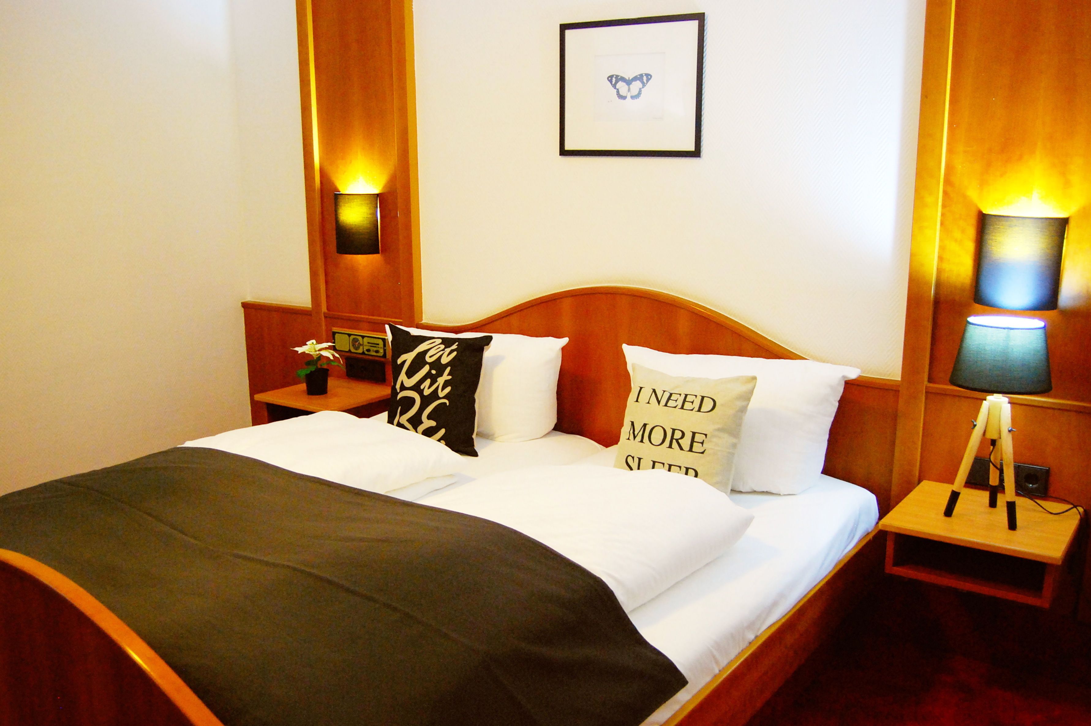 A hotel room with a wooden bed, two bedside tables with lamps, a framed butterfly picture on the wall, and a pillow with the text "I NEED MORE SLEEP".