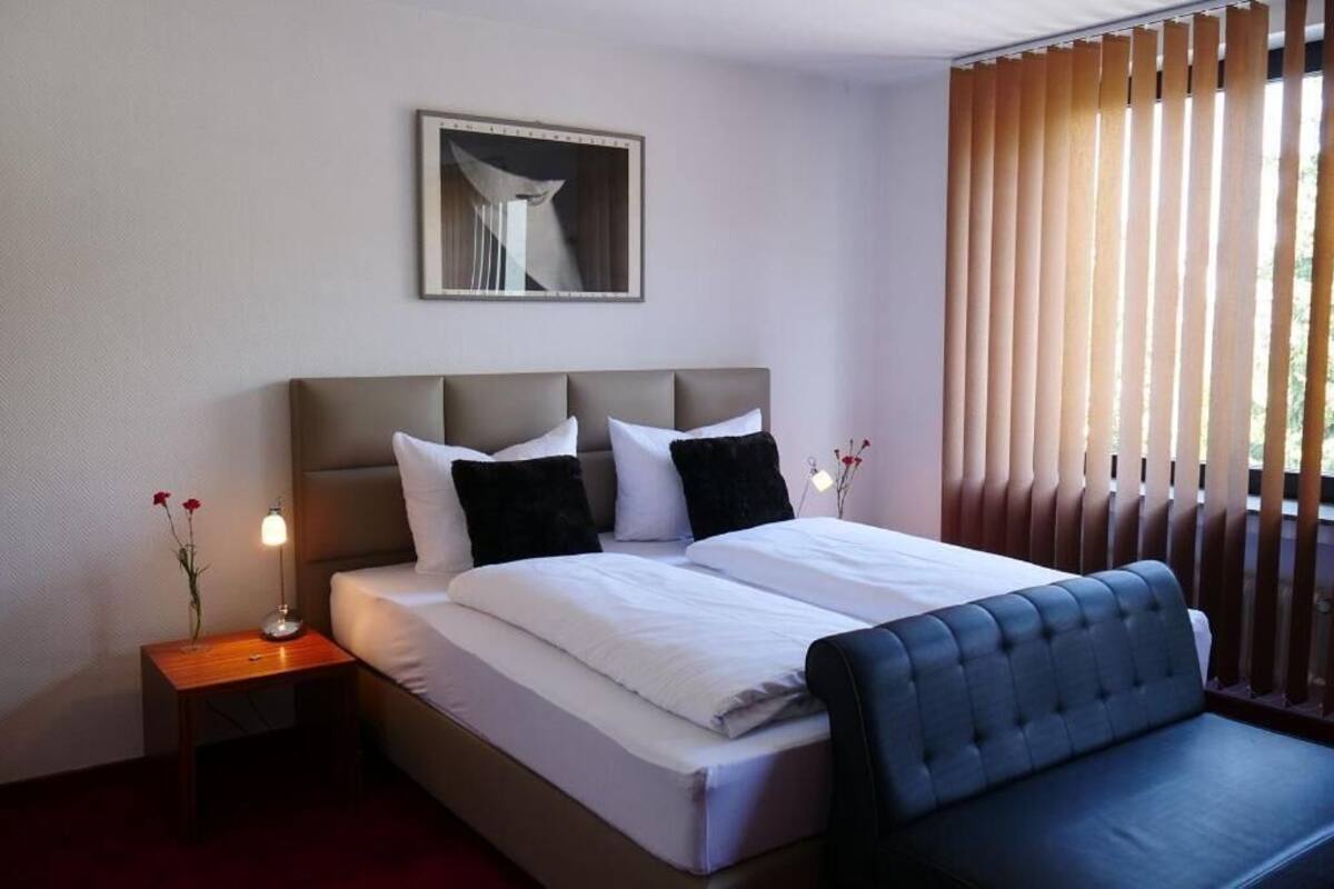 A hotel room with a bed, a bedside table with a lamp, a chair, a window with blinds, and a framed picture on the wall.