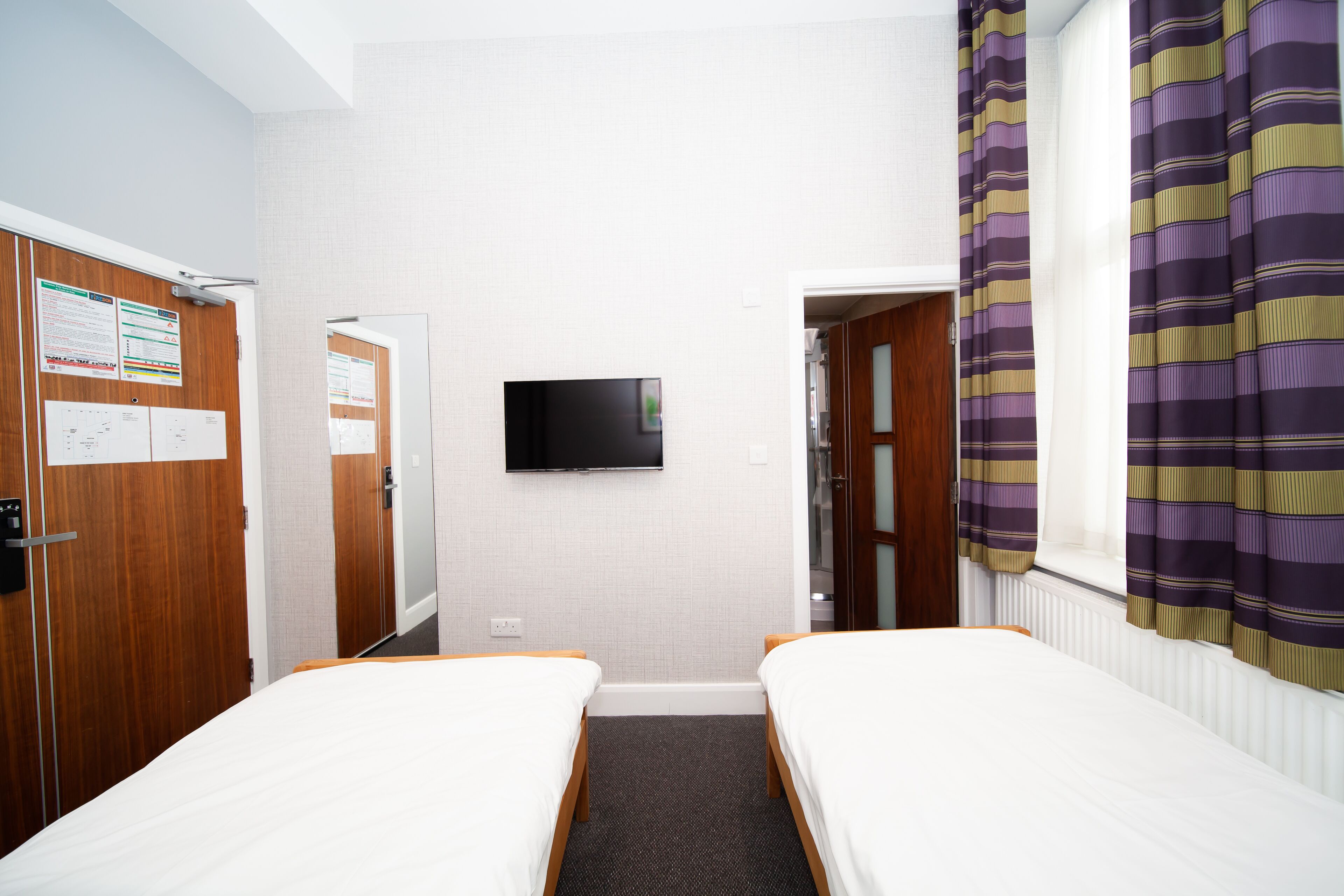 Une chambre d’hôtel avec deux lits simples, un téléviseur à écran plat fixé au mur, des armoires en bois et une fenêtre avec des rideaux à rayures.