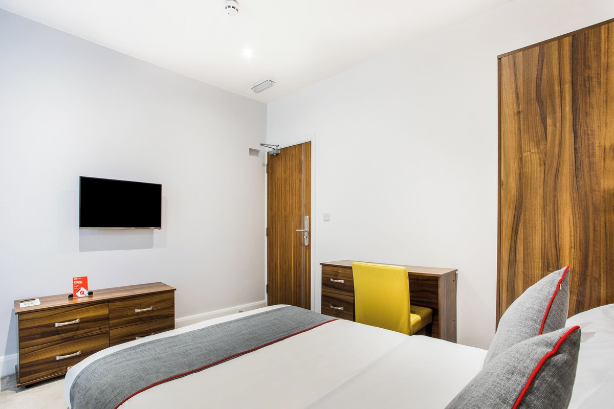 A bedroom with a bed, a wooden wardrobe, a desk with a chair, and a flat-screen TV mounted on the wall.