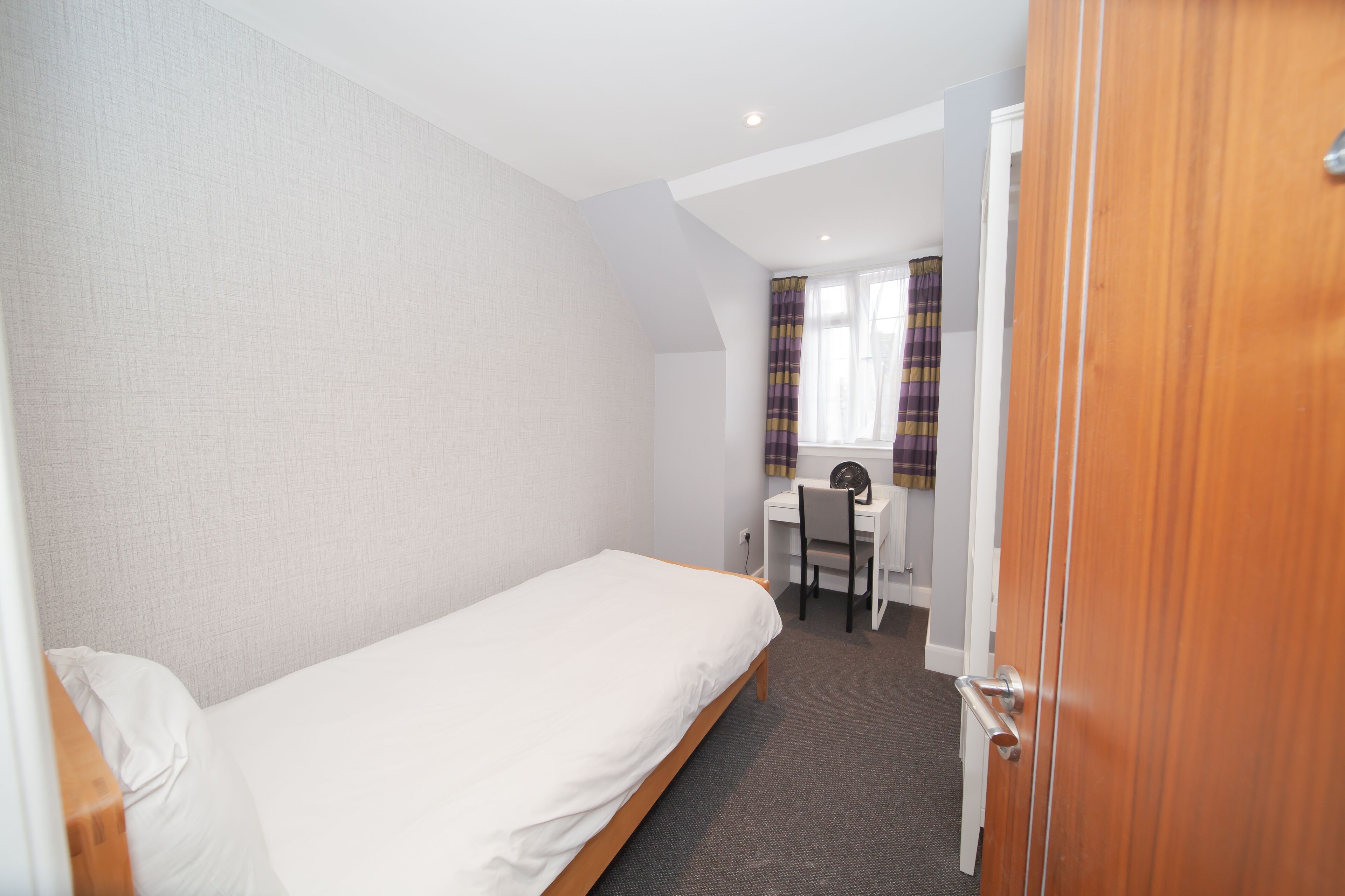 Une petite chambre avec un lit simple, un bureau et une chaise, une fenêtre avec des rideaux et une porte.