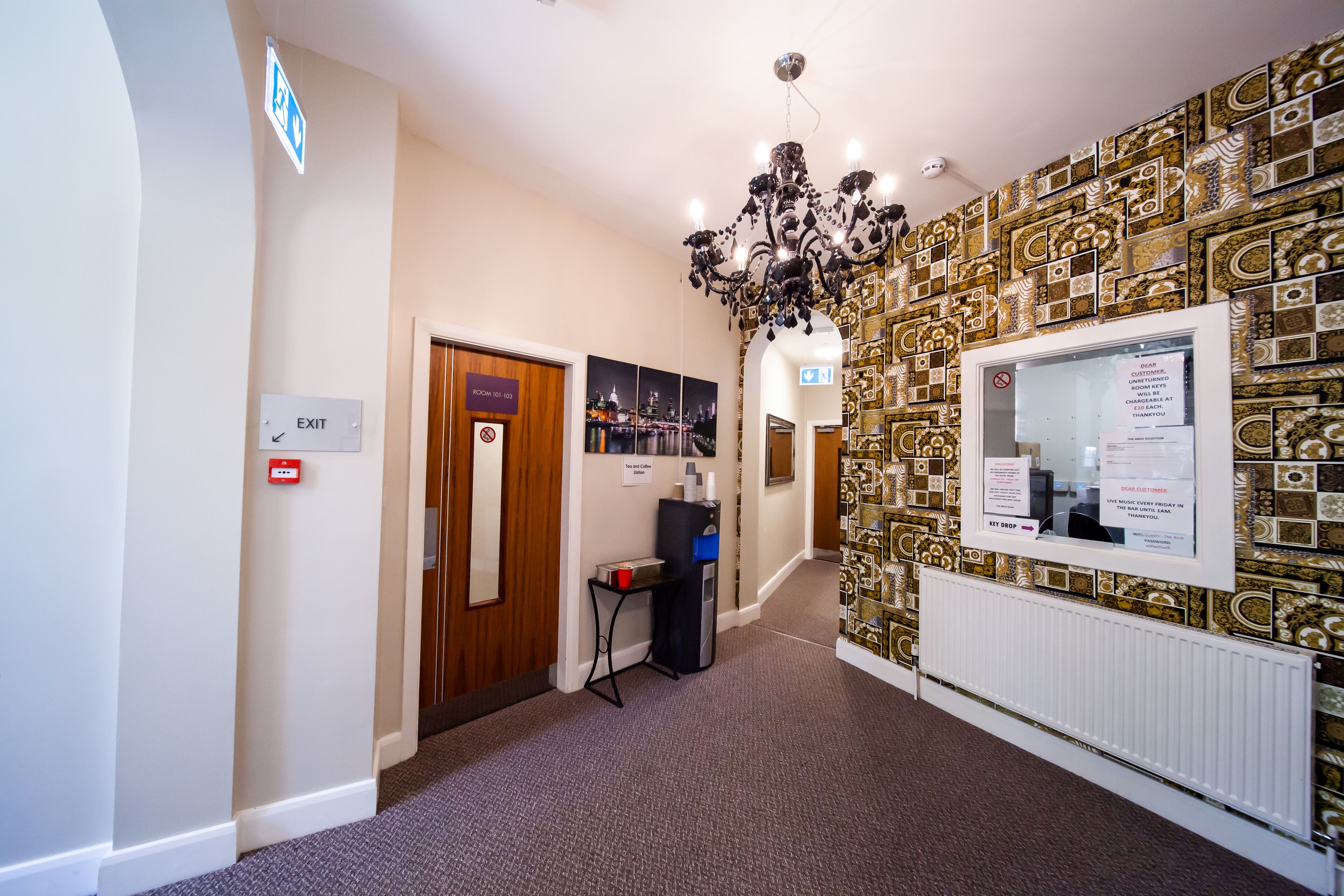 A hotel lobby with an exit sign, a reception desk, a chandelier, and patterned wall tiles.