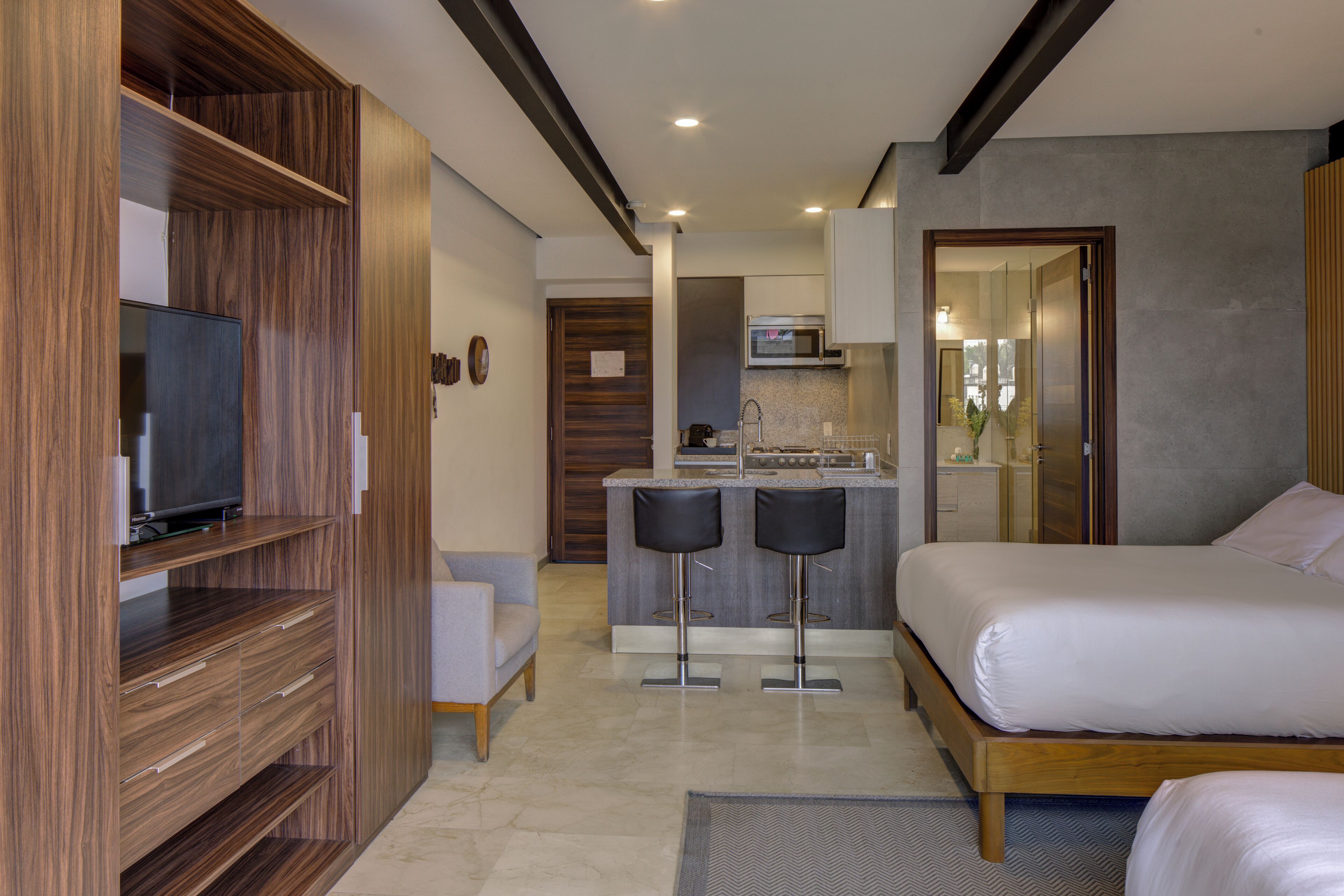 Una habitación de hotel moderna con un armario de madera, televisor, sofá, cama y una cocina pequeña con microondas y un bar.