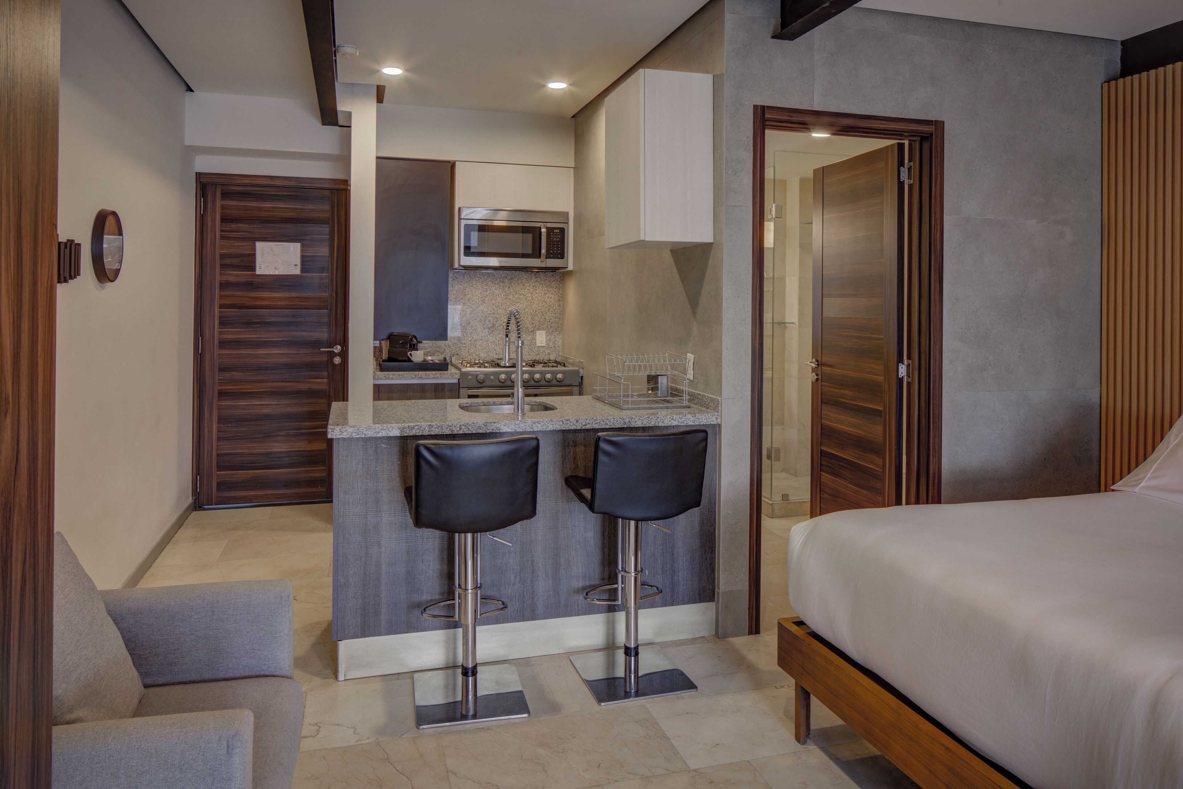 A compact hotel room with a kitchenette, a bed, a small seating area, and a bathroom.
