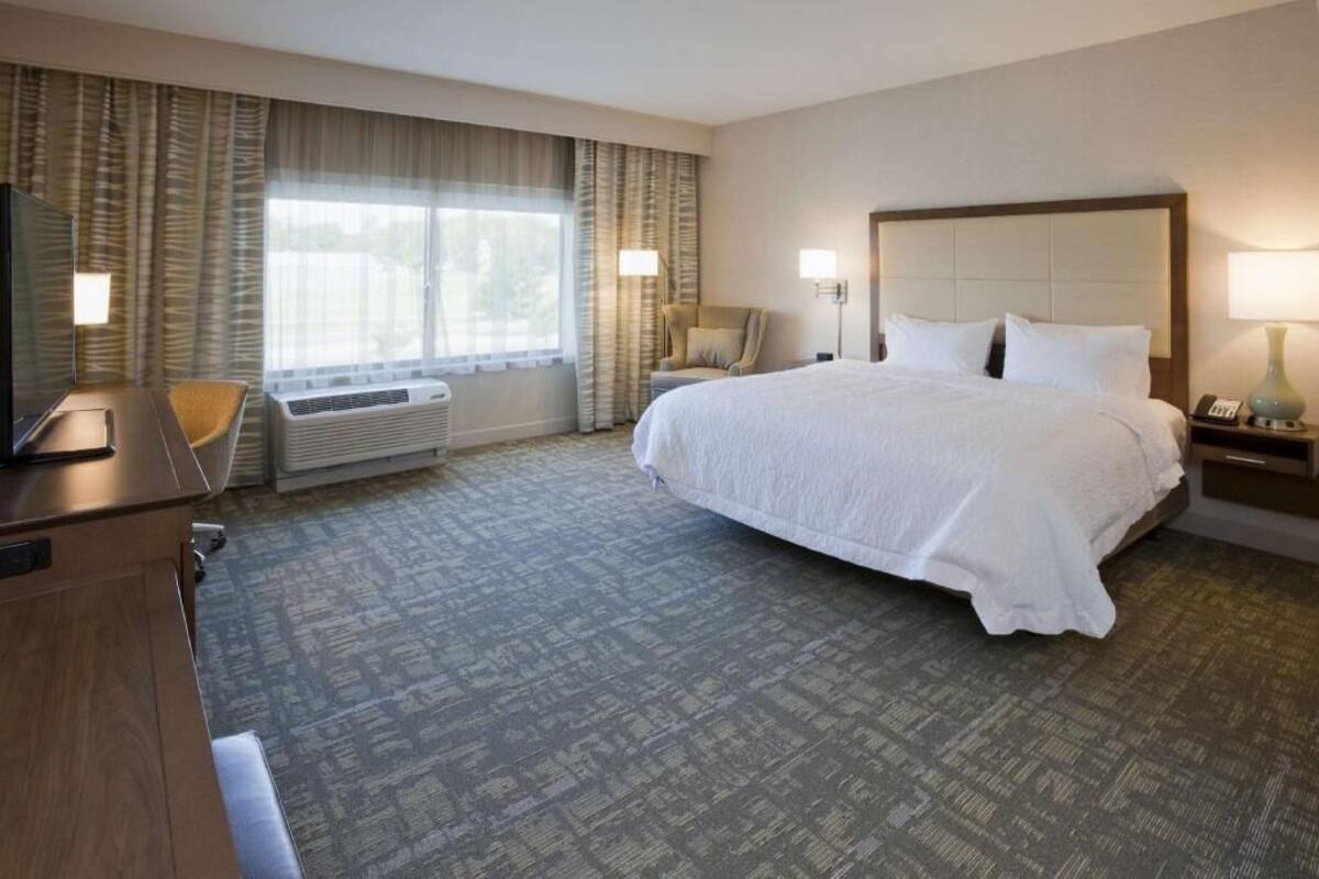 A hotel room with a large bed, a desk, a chair, a window with curtains, and a wall-mounted lamp.