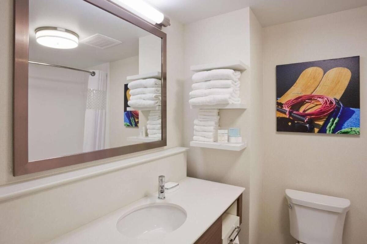 A bathroom with a sink, mirror, toilet, and shelves with folded towels.