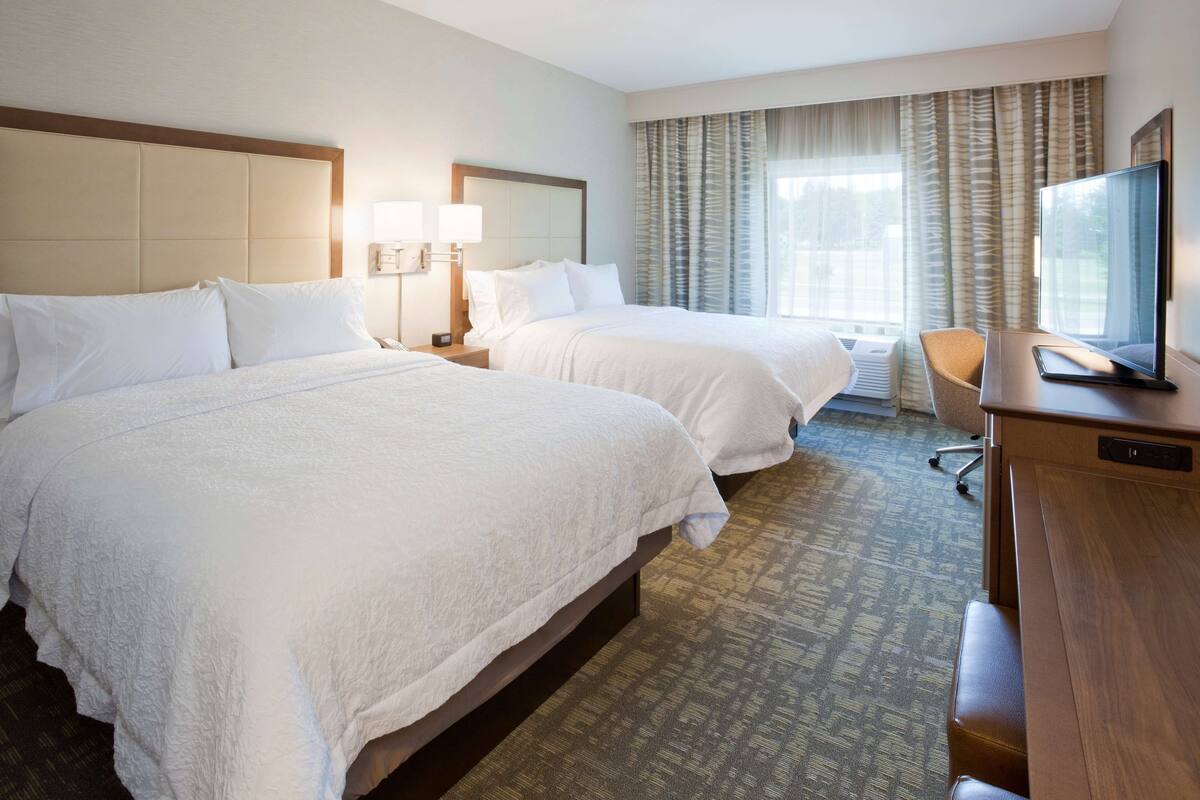 A hotel room with two beds, a desk with a TV, and a window with curtains.