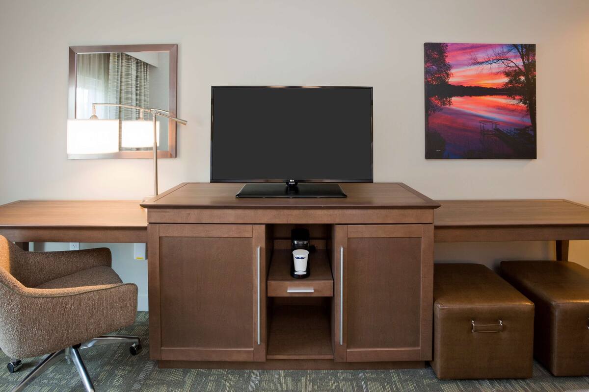 A hotel room with a wooden desk, a flat-screen TV, a lamp, a chair, and a painting on the wall.