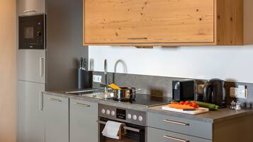 A modern kitchen with grey cabinets, a stainless steel refrigerator, a microwave, a built-in oven, and a wooden upper cabinet.