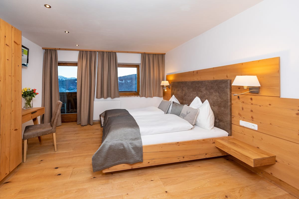A spacious bedroom with a large bed, wooden headboard, bedside tables, a desk, and a view of mountains through the window.