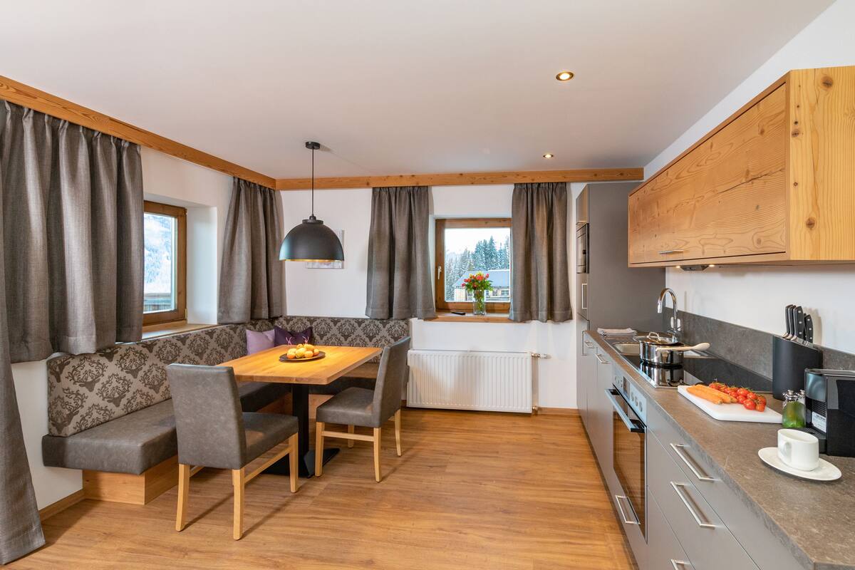 A modern kitchen with a wooden dining table, grey upholstered bench, and a window with curtains.