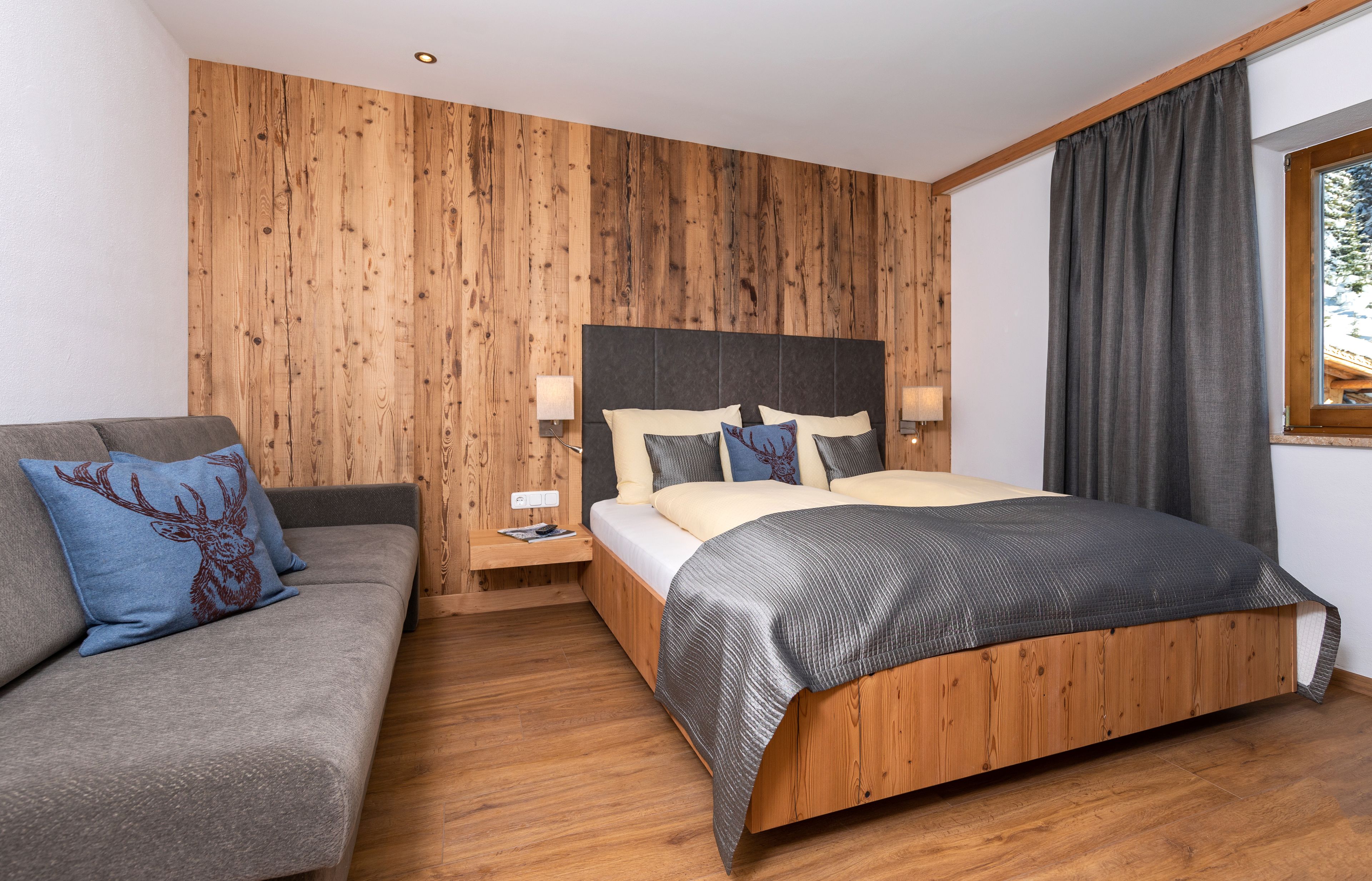 A bedroom with a wooden headboard, a bed with a grey and black bedspread, a grey sofa with a deer pillow, and a nightstand with a lamp.