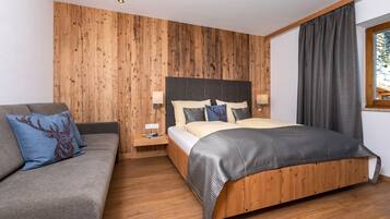 A bedroom with a wooden headboard, a bed with a grey and black bedspread, a grey sofa with a deer pillow, and a nightstand with a lamp.