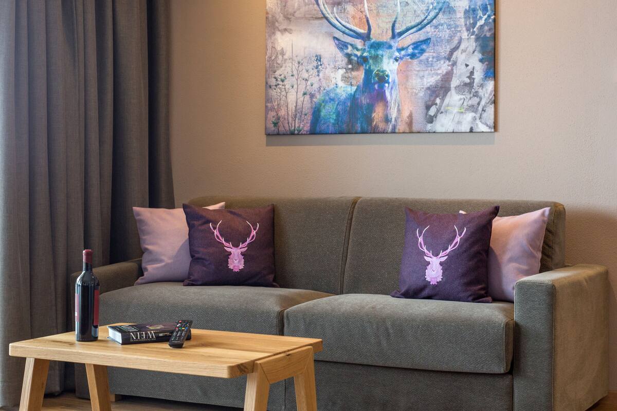 A living room with a grey sofa, two purple pillows with deer designs, a wooden coffee table, a remote control, and a wine bottle.