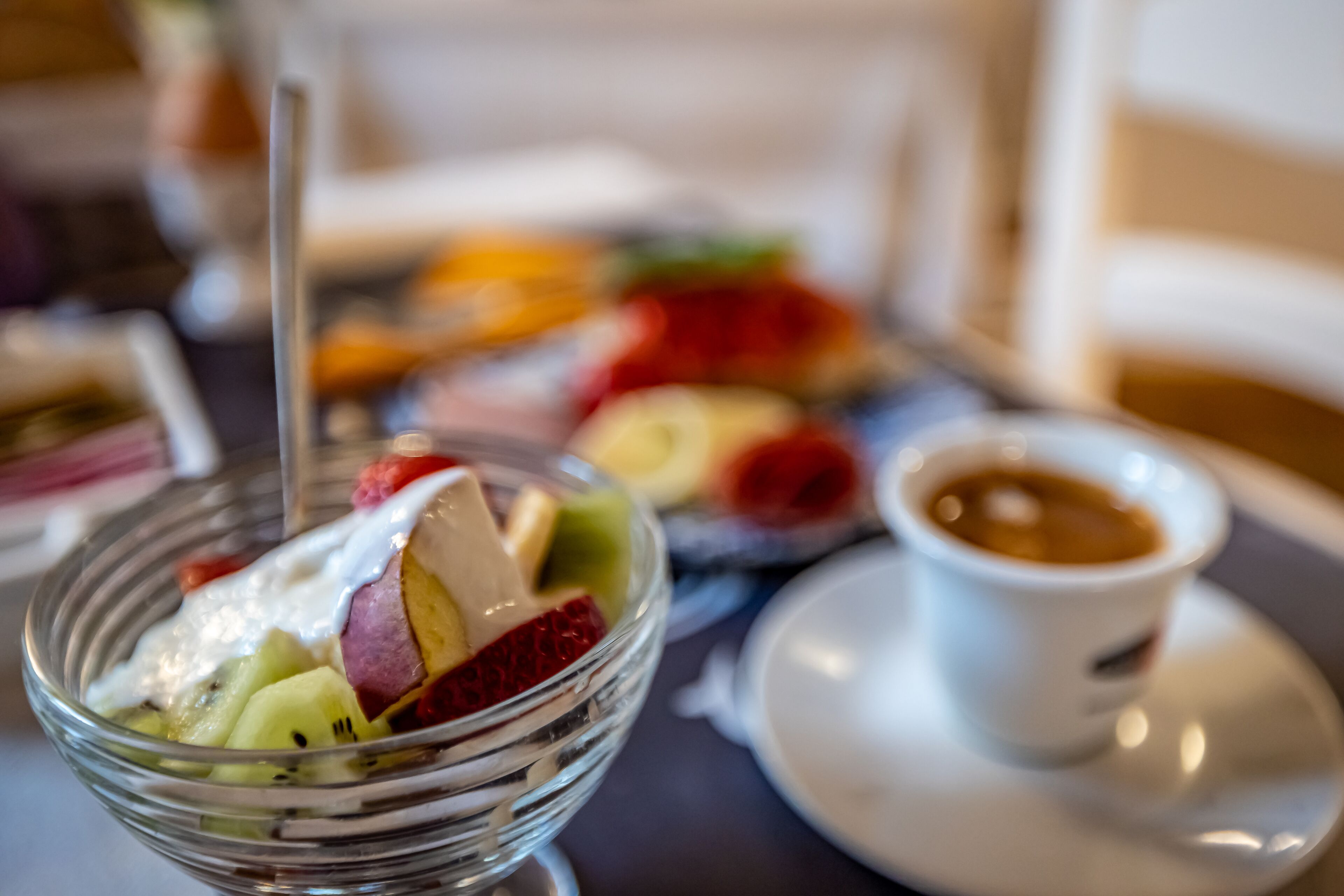 Een glazen schaal met diverse soorten fruit en een lepel, een wit kopje met een schotel, en een wazige achtergrond van een eettafel.