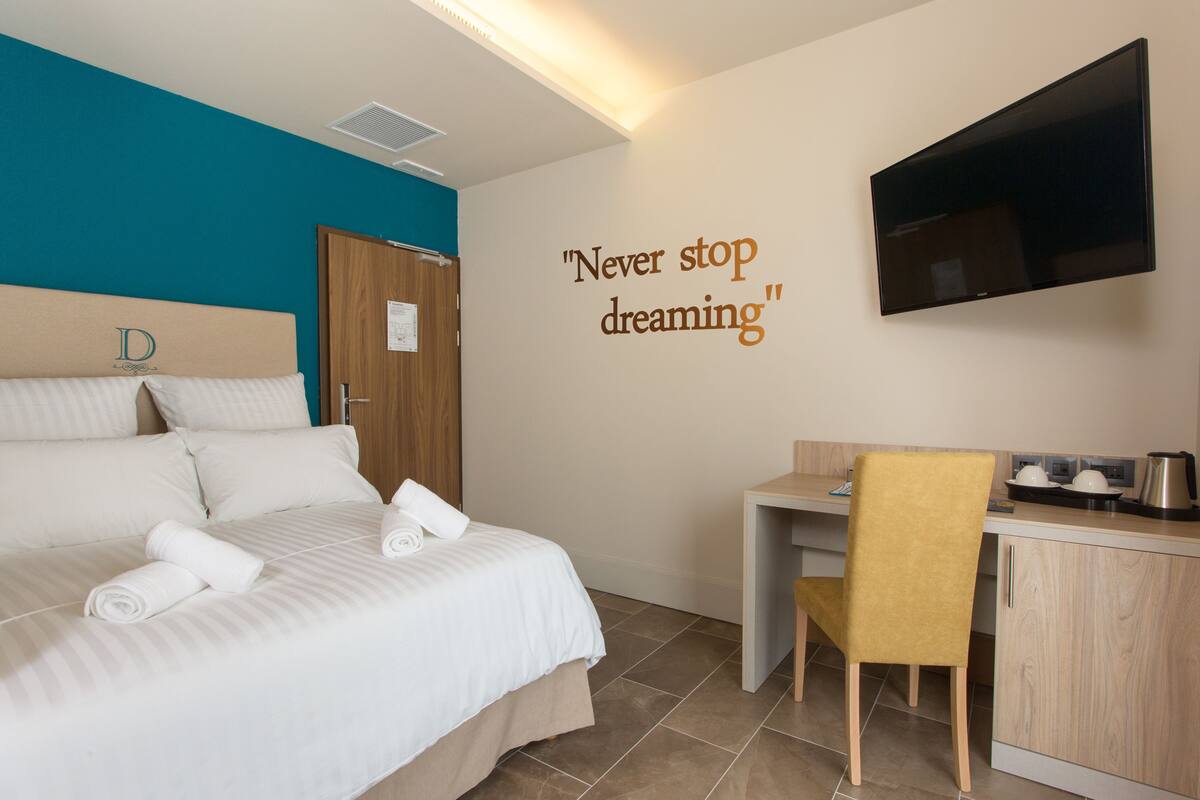 A hotel room with a bed, a desk, a chair, a television, and a motivational quote on the wall.