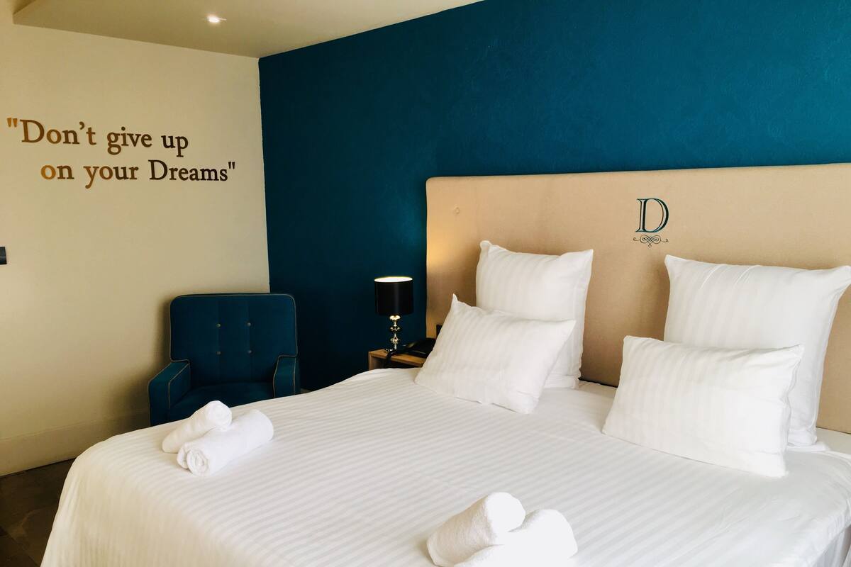 A neatly made bed with white linens and pillows, a blue armchair, a bedside table with a lamp, and a motivational quote on the wall.