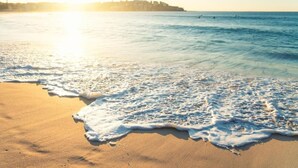 Ein Strand mit klarem Wasser, einem Sandufer und einer entfernten Küstenlinie mit Gebäuden.