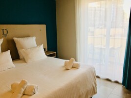 Una cama bien tendida con sábanas y almohadas blancas, un cabecero de madera con la letra 'D', una mesita de noche pequeña y una ventana con cortinas traslúcidas.
