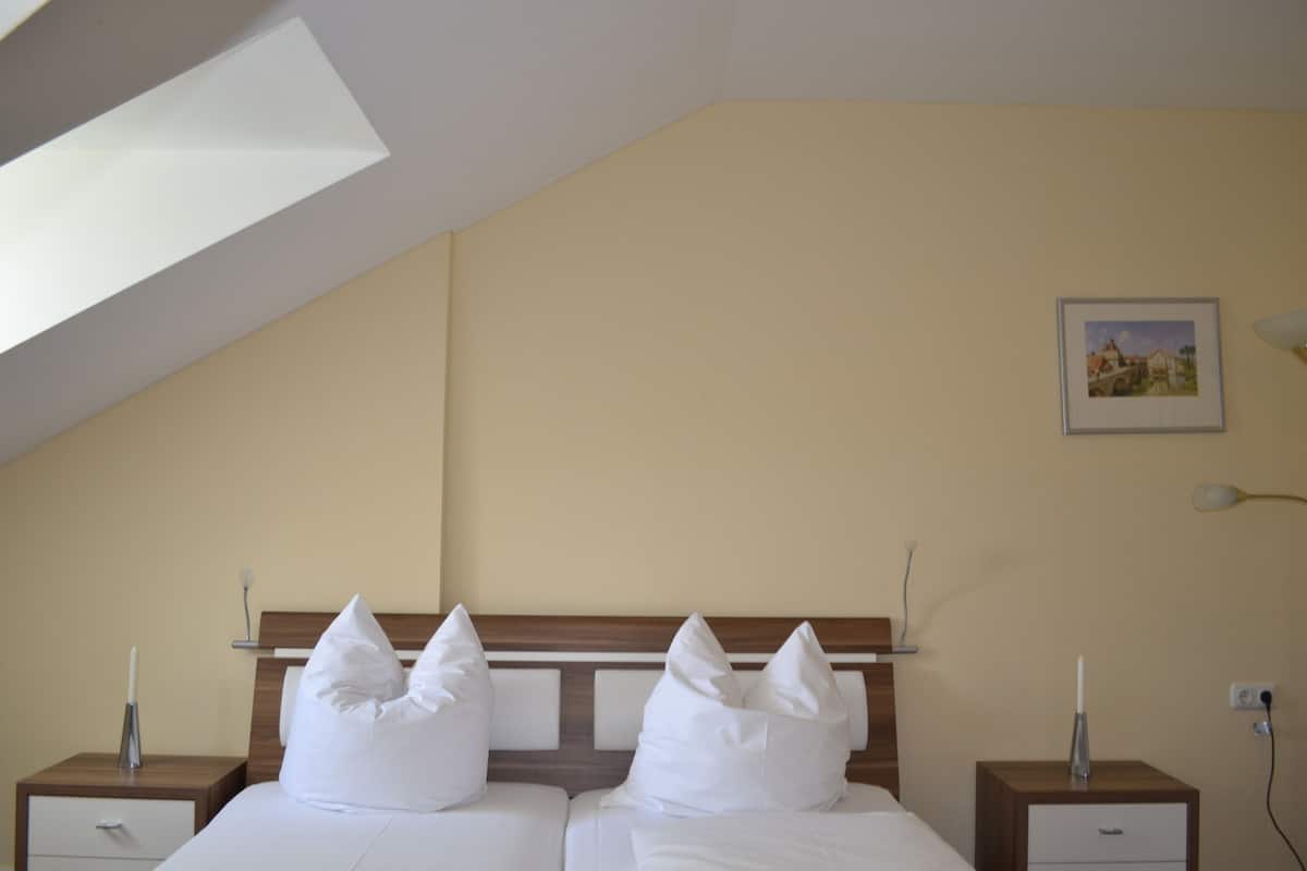 A bedroom with a sloped ceiling, a wooden headboard, two beds with white linens, bedside tables with lamps, and a framed picture on the wall.