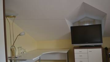 Una habitación con un escritorio blanco, un televisor de pantalla plana sobre un mueble de madera y una lámpara de pie.