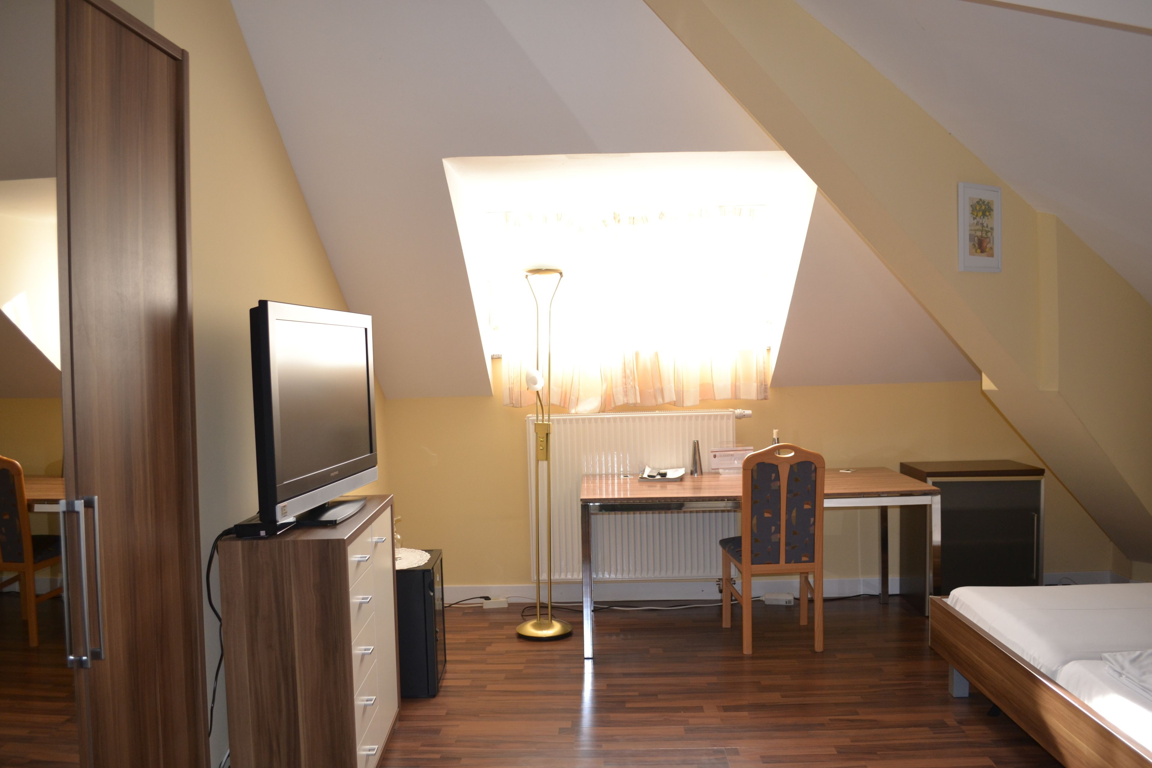 Una habitación con techo inclinado, un escritorio con silla, una cama, un televisor sobre un mueble de madera y un radiador.