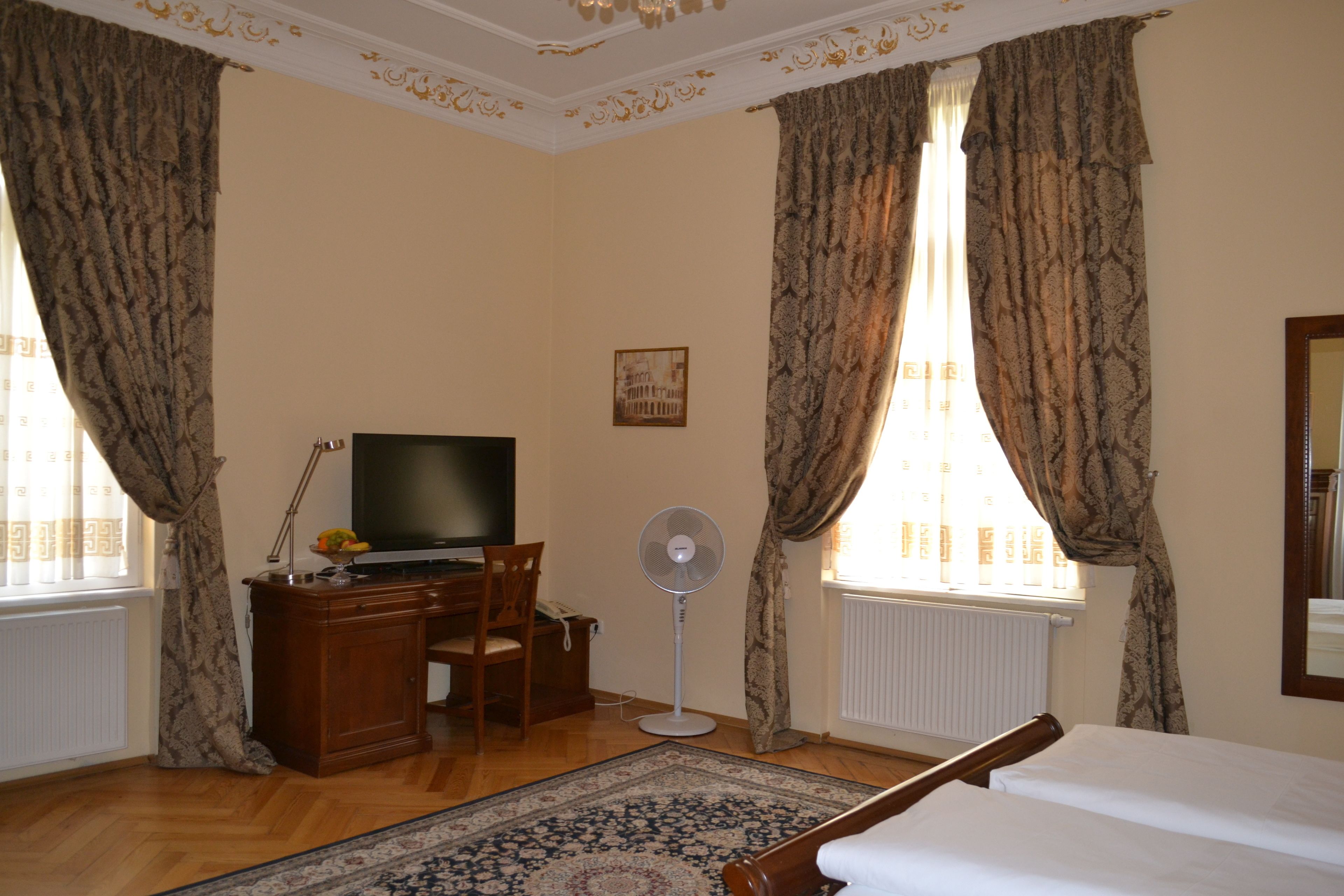 Una habitación de hotel con un escritorio de madera, un televisor, un ventilador de pie, una cama y ventanas grandes con cortinas.