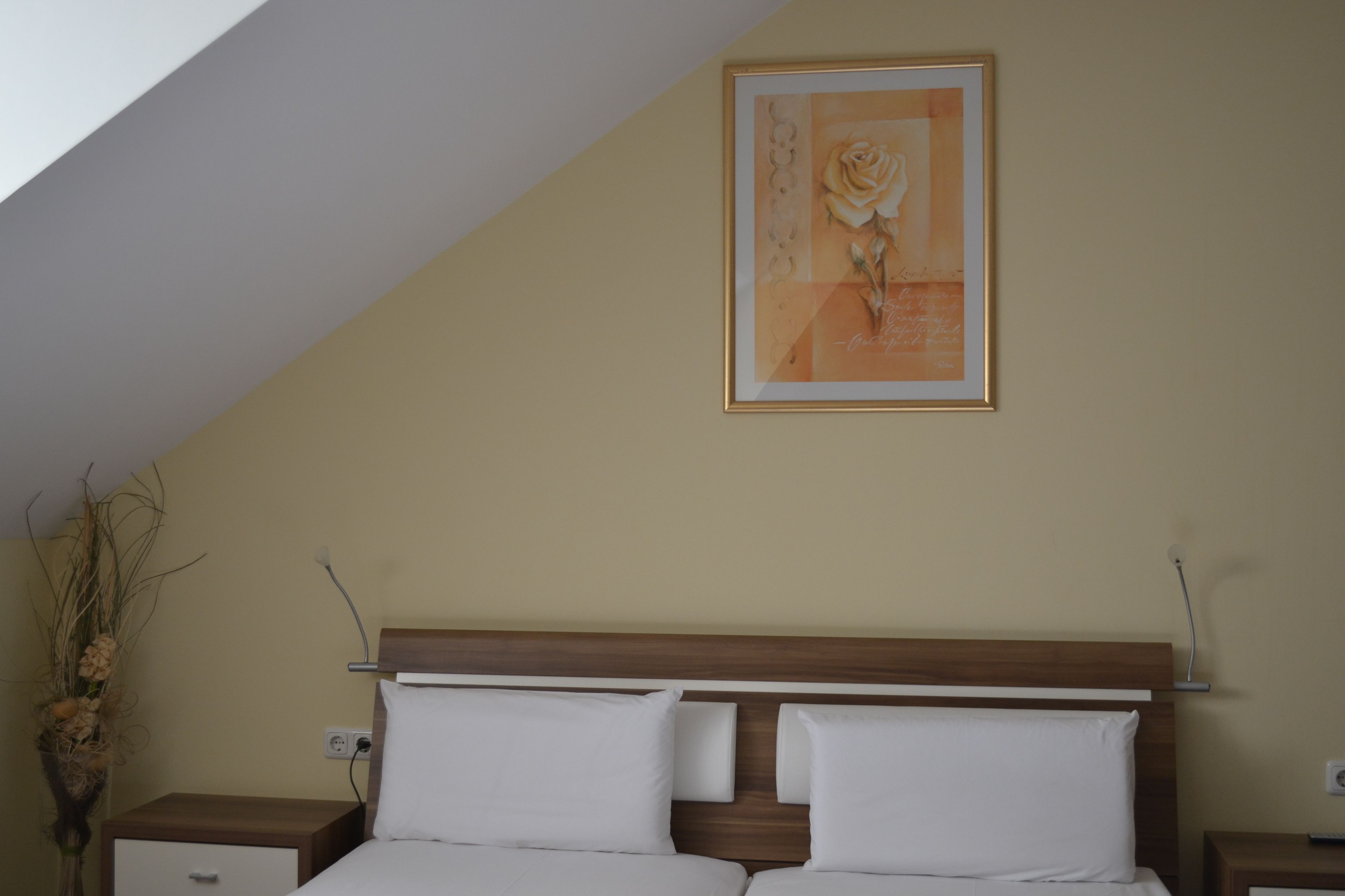 A bedroom with a wooden headboard, two white pillows, a framed artwork of a rose, and a nightstand with a vase of dried plants.
