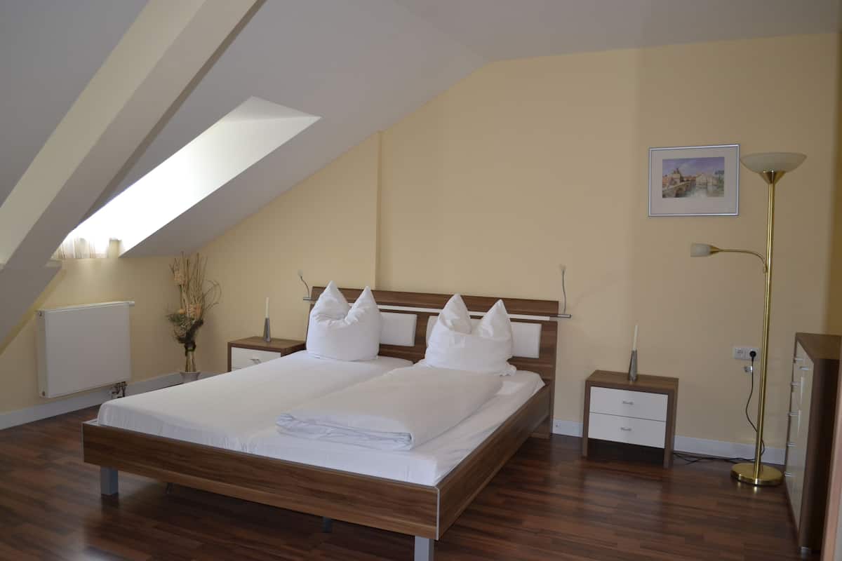 A bedroom with a sloped ceiling, a bed with white bedding, a bedside table, a floor lamp, and a picture on the wall.
