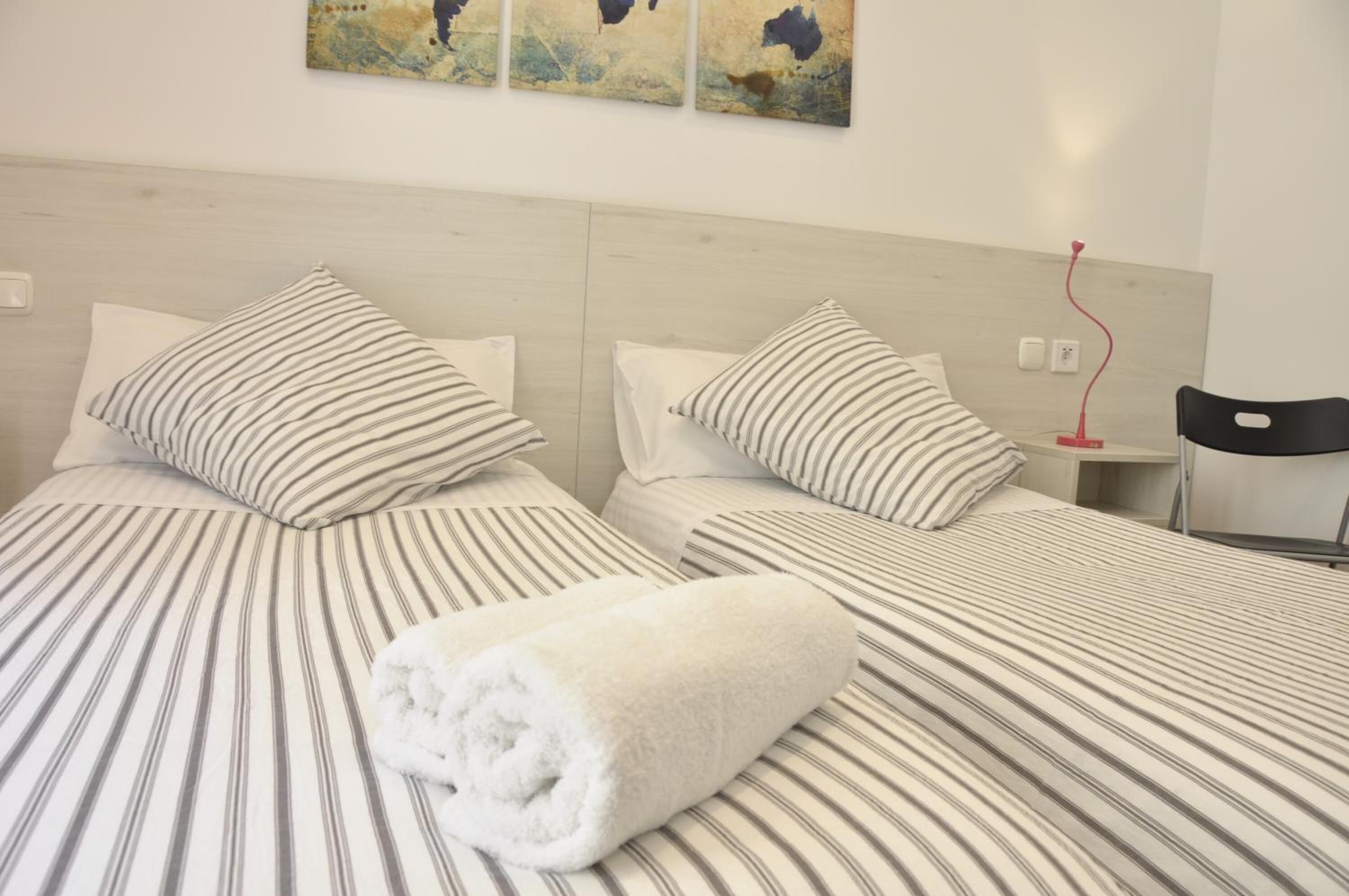 Une chambre d’hôtel avec deux lits, une literie à rayures, une serviette blanche roulée, une table de chevet avec une lampe rouge et des œuvres d’art abstraites accrochées au mur.