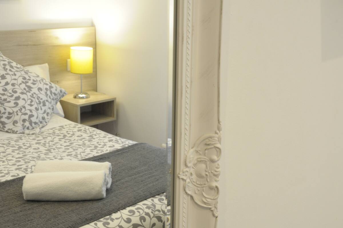 Een net opgemaakt bed met behangpatroon, een bedlampje en een opgevouwen handdoek op de vloer.