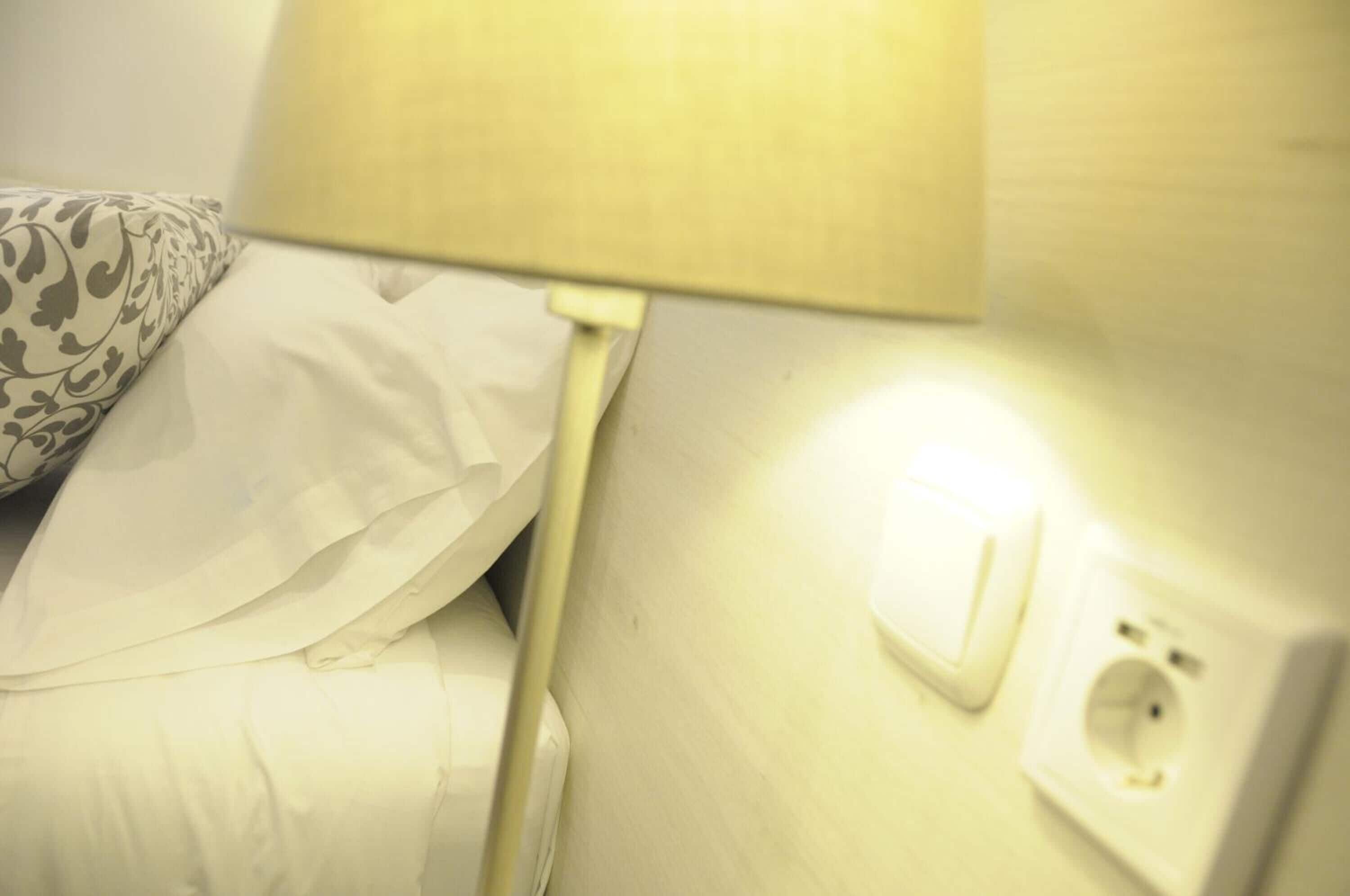 Ein Bett mit weißer Bettwäsche und einem gemusterten Kopfkissen, ein Bettlampenschalter und eine Steckdose an der Wand.