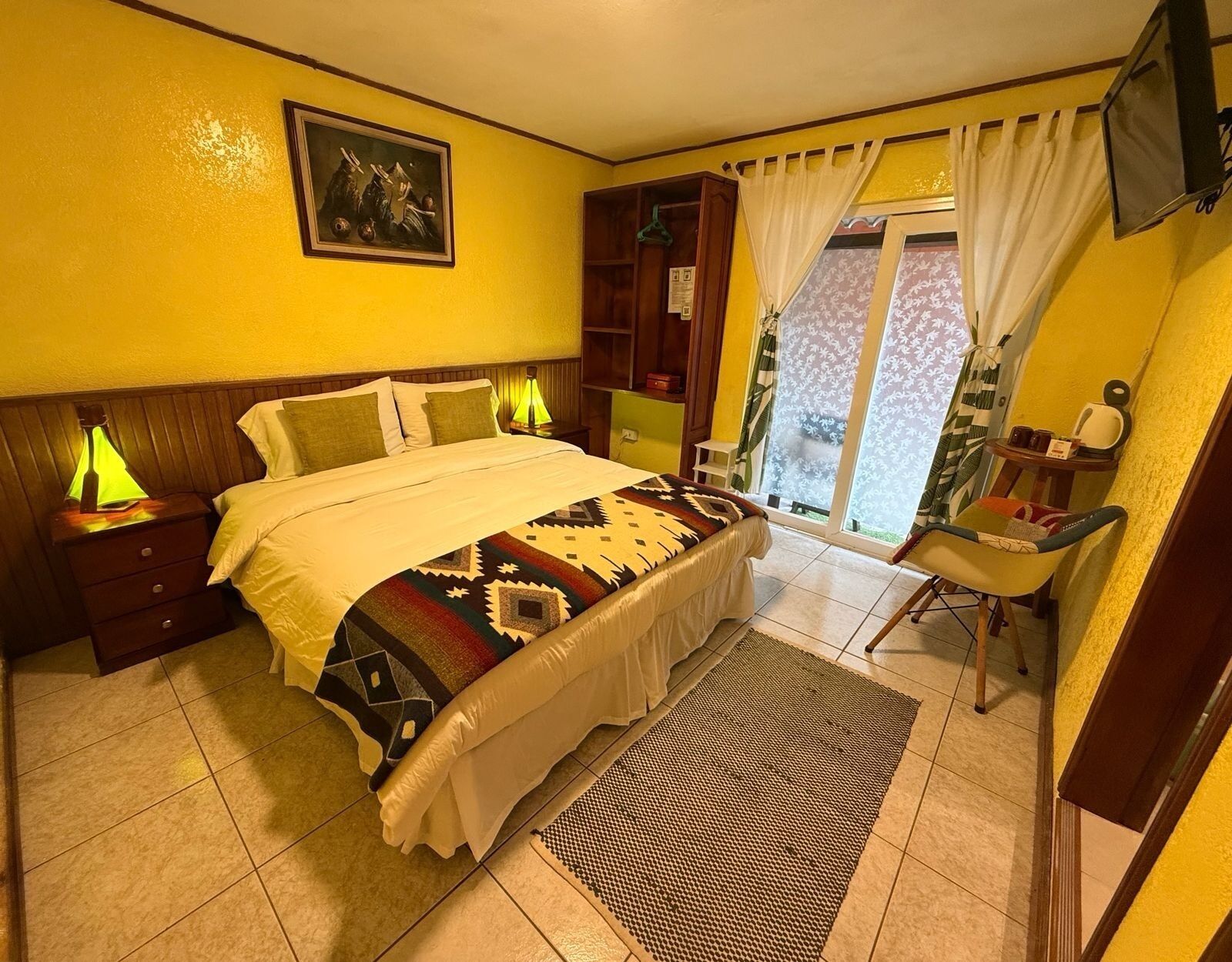 A bedroom with a bed, bedside lamps, a chair, a desk, a window with curtains, and a painting on the wall.