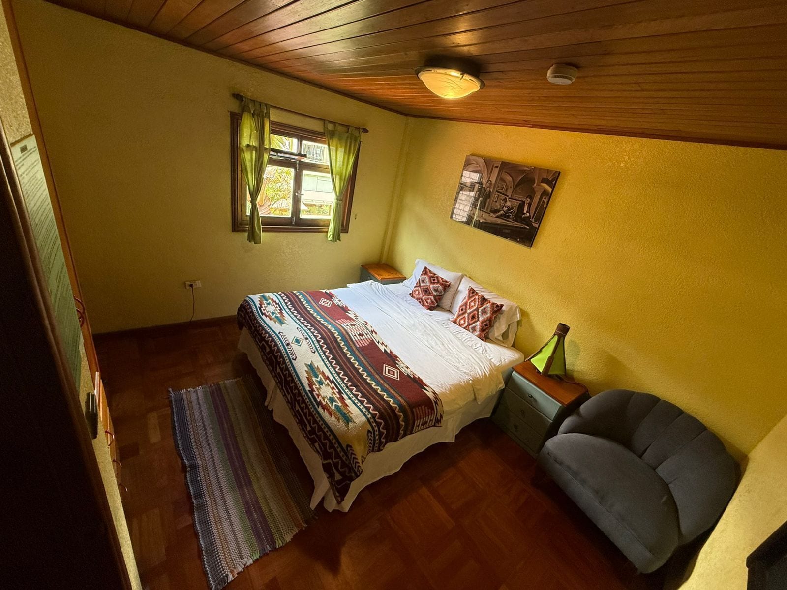 A bedroom with a wooden ceiling, a bed with patterned bedding, a nightstand, a chair, and a window with curtains.