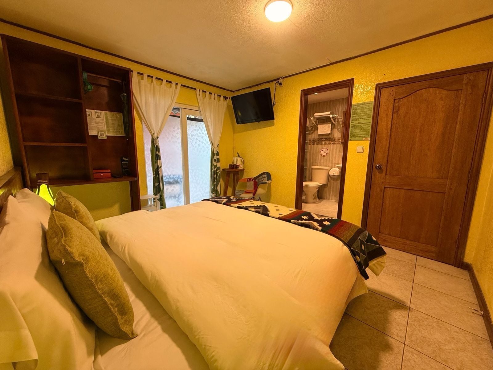 A bedroom with a bed, a television mounted on the wall, a wooden door, and a small table with a lamp.
