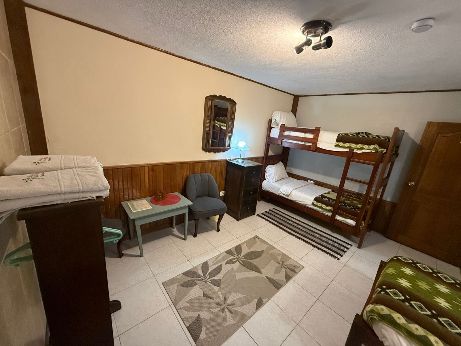 Una habitación pequeña y funcional con una cama litera, un escritorio con silla, una mesita, un espejo y una puerta.