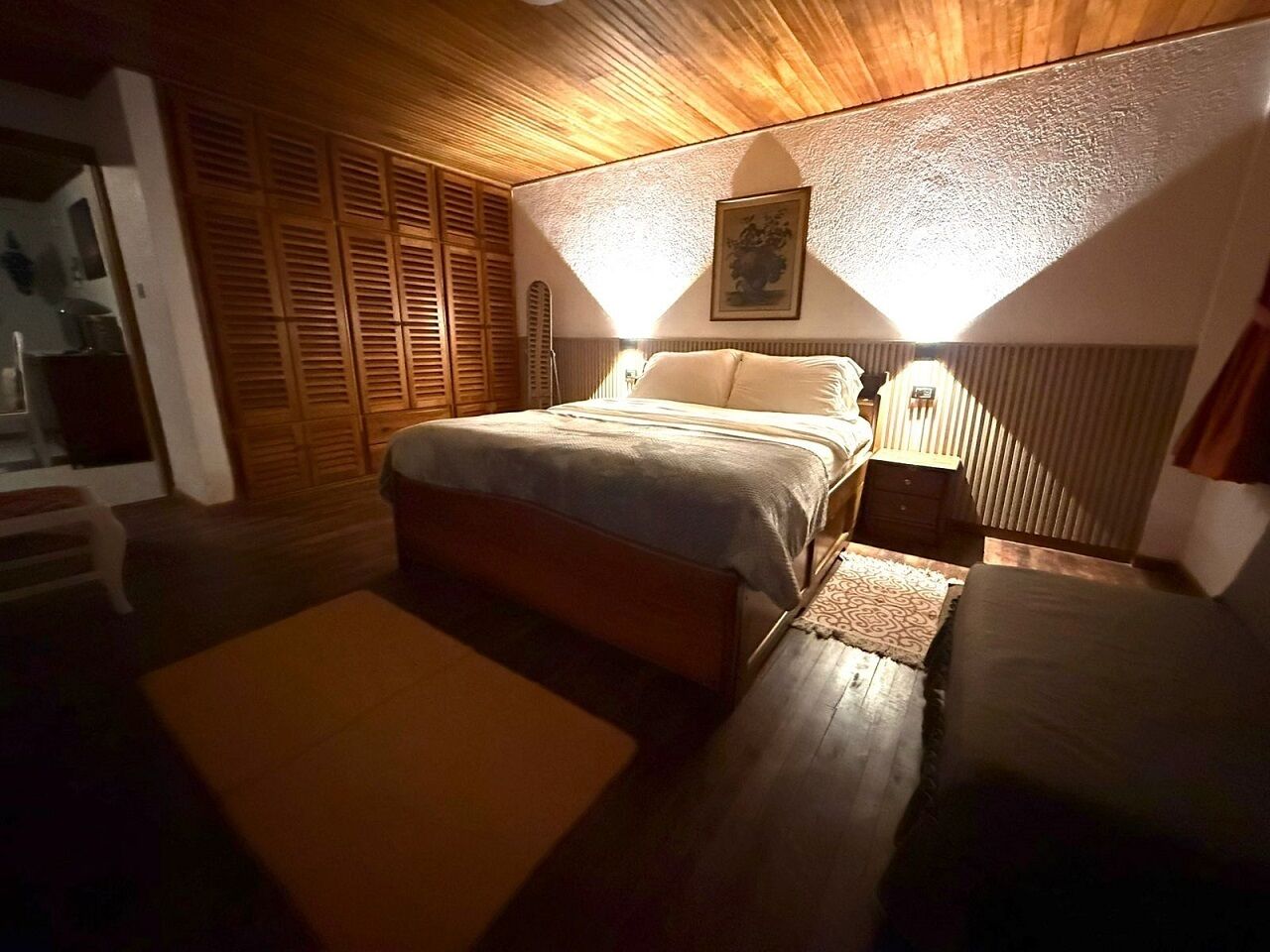 Un dormitorio con cama, mesitas de noche, un armario de madera y una pequeña zona de estar.