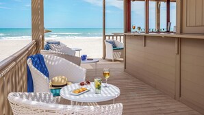 Un balcón frente a la playa con muebles de mimbre, una mesa con bebidas y vistas al océano.