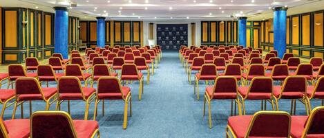 A conference room with rows of red chairs and blue carpet, featuring blue columns and wooden panel walls.