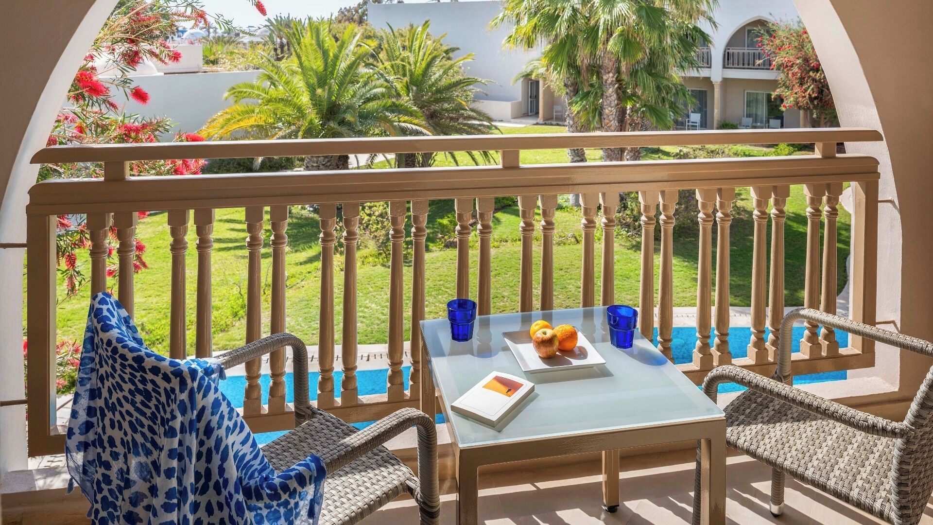 En balkon med fletmøbler, et glasbord og udsigt til en pool og grønt område.