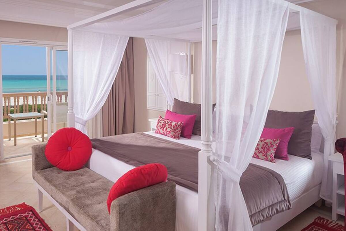 A bedroom with a four-poster bed, a bench, a red chair, and a view of the beach.